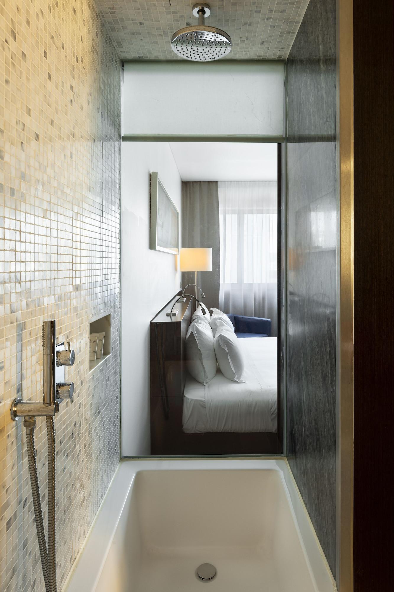 Vip Grand Lisboa Hotel & Spa Exterior photo The photo shows a modern hotel room viewed through an entrance that appears to lead from a bathroom area. On the left, there is a tiled wall, likely from a shower or bathroom. In the main room, there is a bed with white linens and multiple pillows, a