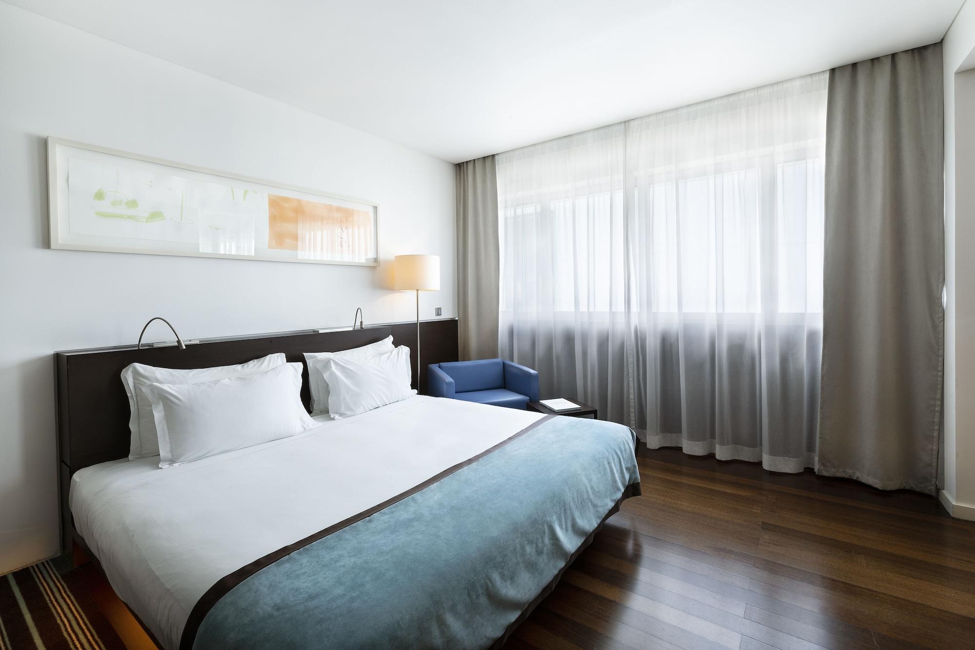 Vip Grand Lisboa Hotel & Spa Exterior photo The photo shows a modern hotel room featuring a large bed with white sheets and a light blue blanket. There are two pillows on the bed. Next to the bed, there’s a small blue chair and a window covered with sheer curtains that allow natural light to e